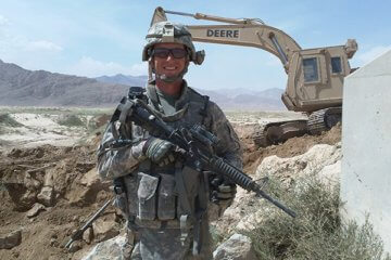 El soldado del ejército estadounidense Brian Ross sosteniendo un rifle en un sitio de construcción en Afganistán con una retroexcavadora de fondo.