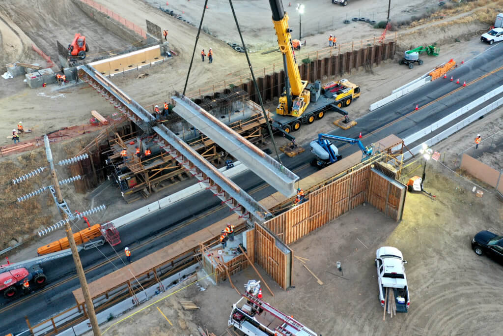 A shot from above, showing a concrete girder being placed on columns and abutments by a crane over state route 46 at dusk. Other heavy machinery and workers and vehicles also visible.