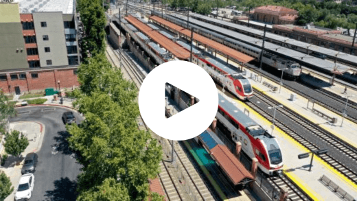 Image of a train station. There are 8 tracks visible. There are two sleek, white and red trains at the station. To the right of those trains is an older looking grey train. All of the trains have Caltrain logos on them. To the left of the train station is a street lined with trees and three cars. A white sedan and a grey sedan are parked and a grey sedan is driving. Behind the street is an apartment complex. In the center of the image is a play button, which consists of a white circle with a transparent sideways triangle in the middle.