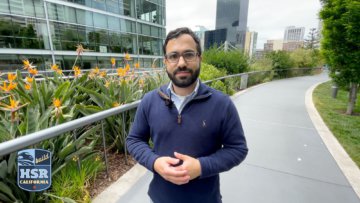 Man walking along a pathway with flowers to the left and grass and trees to the right. The man is wearing glasses, and a blue quarter zip with a checkered button up shirt and white undershirt. The man has short dark hair and beard. In the background are tall buildings and skyscrapers. There is a blue and light blue logo in the bottom left corner that reads 