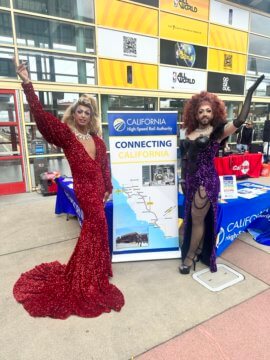 Two people standing next to a banner. The person on the left has a full face of make-up, long blonde hair, a big silver necklace, and a long sparkly red dress. The person on the right is wearing a sparkly black and purple dress, black stockings with black high heels, and elbow length black party gloves. The person on the left has large ginger hair, a full face of make-up, and a dark beard. The banner reads 