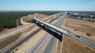 Aerial image of a concrete overpass spanning over railroad tracks and opposing lanes of a highway. The tracks and highway are separated by patches of dirt. There are cars on the highway and about to cross the overpass. In the background on the left is farmland, and on the right is housing.