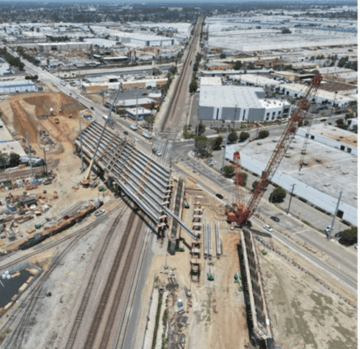 Aerial view of construction on a major intersection. The intersection features four roads feeding into it with railroads also crossing through it. There is visible signs of construction with large pillars being set and a huge red crane nearby. Businesses and parking lots surround the intersection.