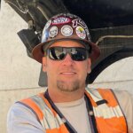 A man wearing a hard hat covered in stickers, sunglasses, a beige shirt, and an orange safety vest smiles at the camera.