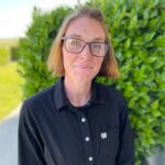 A person with blonde hair, chin length hair and glasses, wearing a black shirt smiles at the camera in front of a bush.