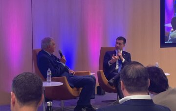 U.S. Transportation Secretary Pete Buttigieg sitting in a chair next to former U.S. Transportation Secretary Ray LaHood. Both men are in suits holding microphones.