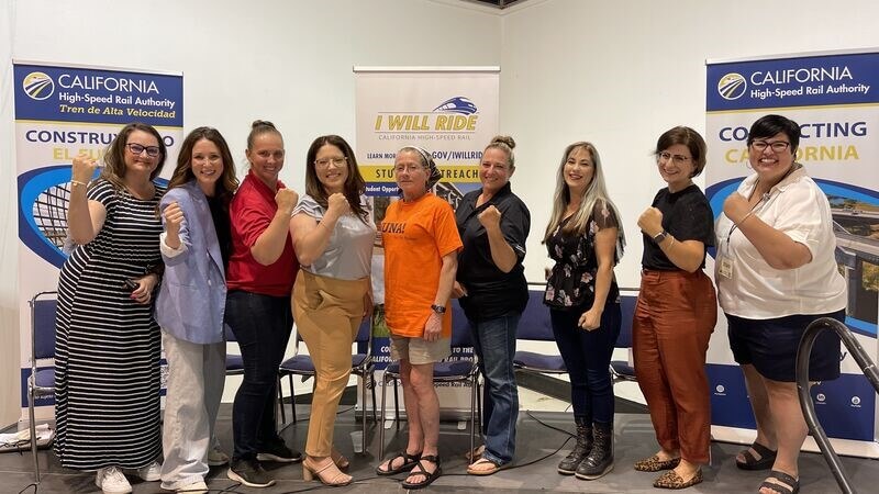 Nine women on a stage smiling and doing a hand muscle flex.