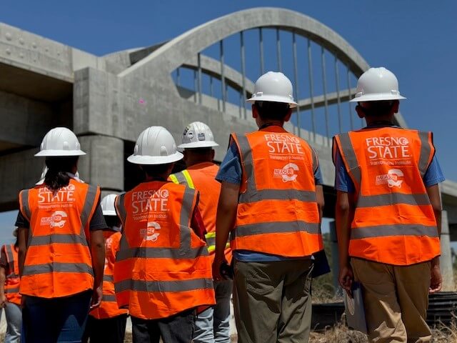 Four students in construction vests that have Fresno State and Measure C logos looking at a large high-speed rail structure. 