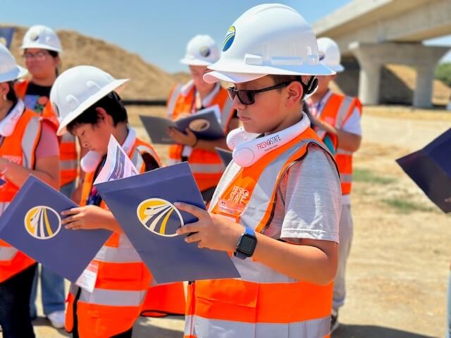 Young kid in a construction vest and hard hat reading something in a folder.