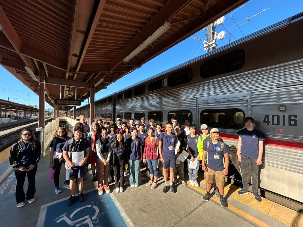 Large group of students in matching shirts that stand on a platform with the Caltrain behind them.