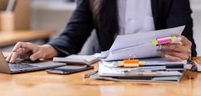 A woman sitting at a desk and reading through papers and working on her computer. Only the woman's hands and torso are in frame.